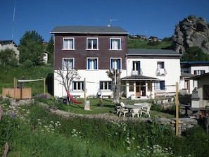A two-story house with a garden, outdoor seating, and a view of a rocky hill.