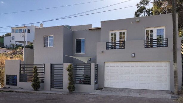 A modern two-story house with a garage, a balcony, and a small tree in the front yard. - Casa Flores & Flores (Ensenada)
