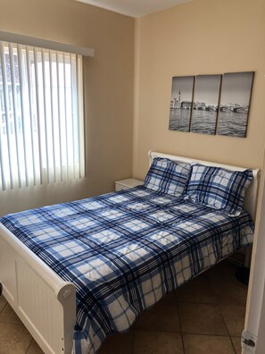 Une chambre à coucher comprenant un lit, une table de chevet, une fenêtre avec des stores et un mur orné de trois tableaux encadrés.