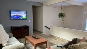 A living room with a television displaying a sports event, a wooden coffee table, and a beige sofa with patterned cushions. - Casa Flores & Flores (Ensenada)