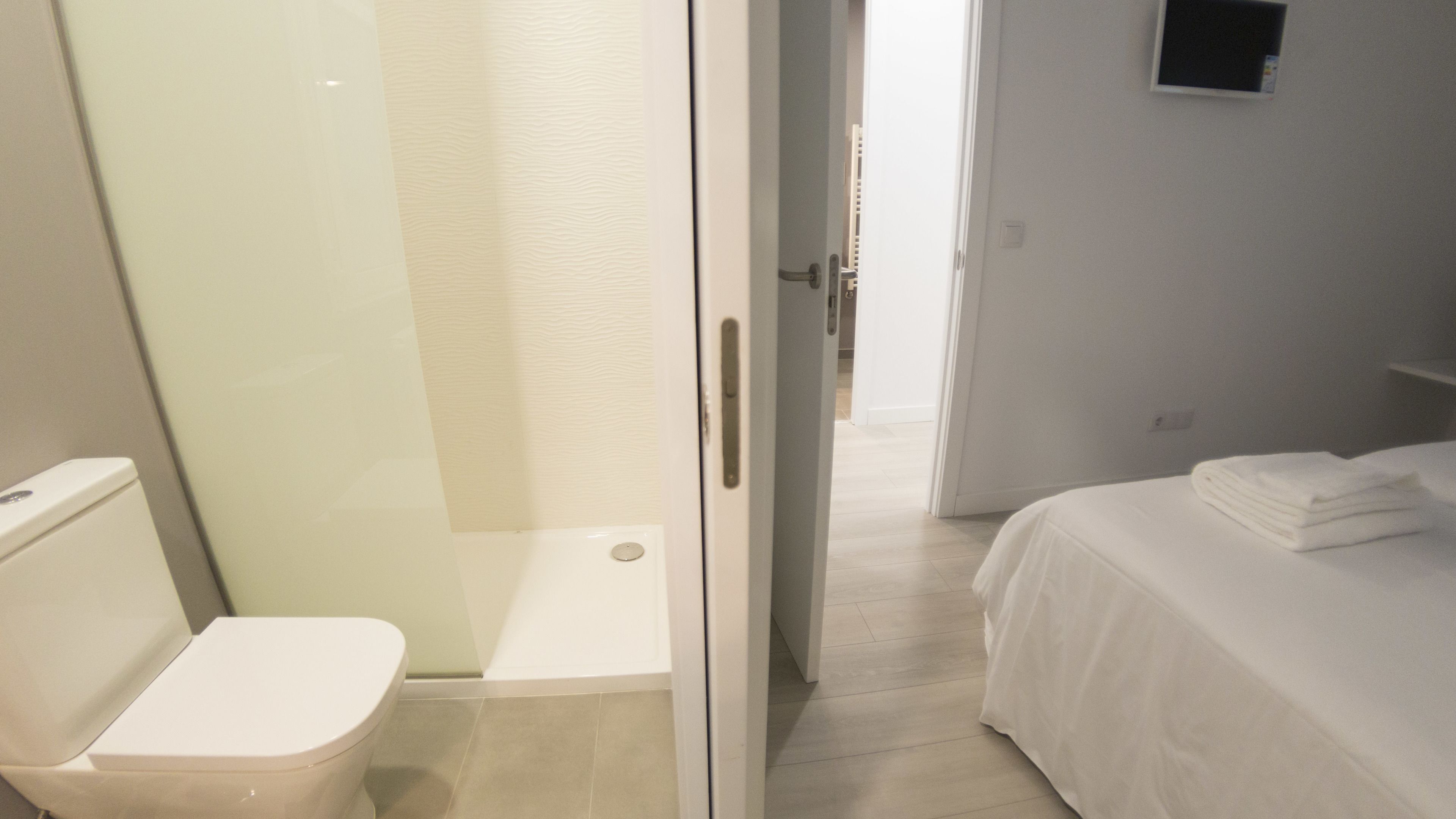 A hotel room with a toilet, a glass shower enclosure, a bed with a neatly folded white towel, and a wall-mounted TV.
