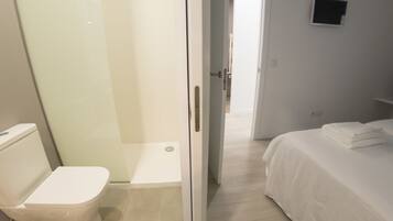 A hotel room with a toilet, a glass shower enclosure, a bed with a neatly folded white towel, and a wall-mounted TV.