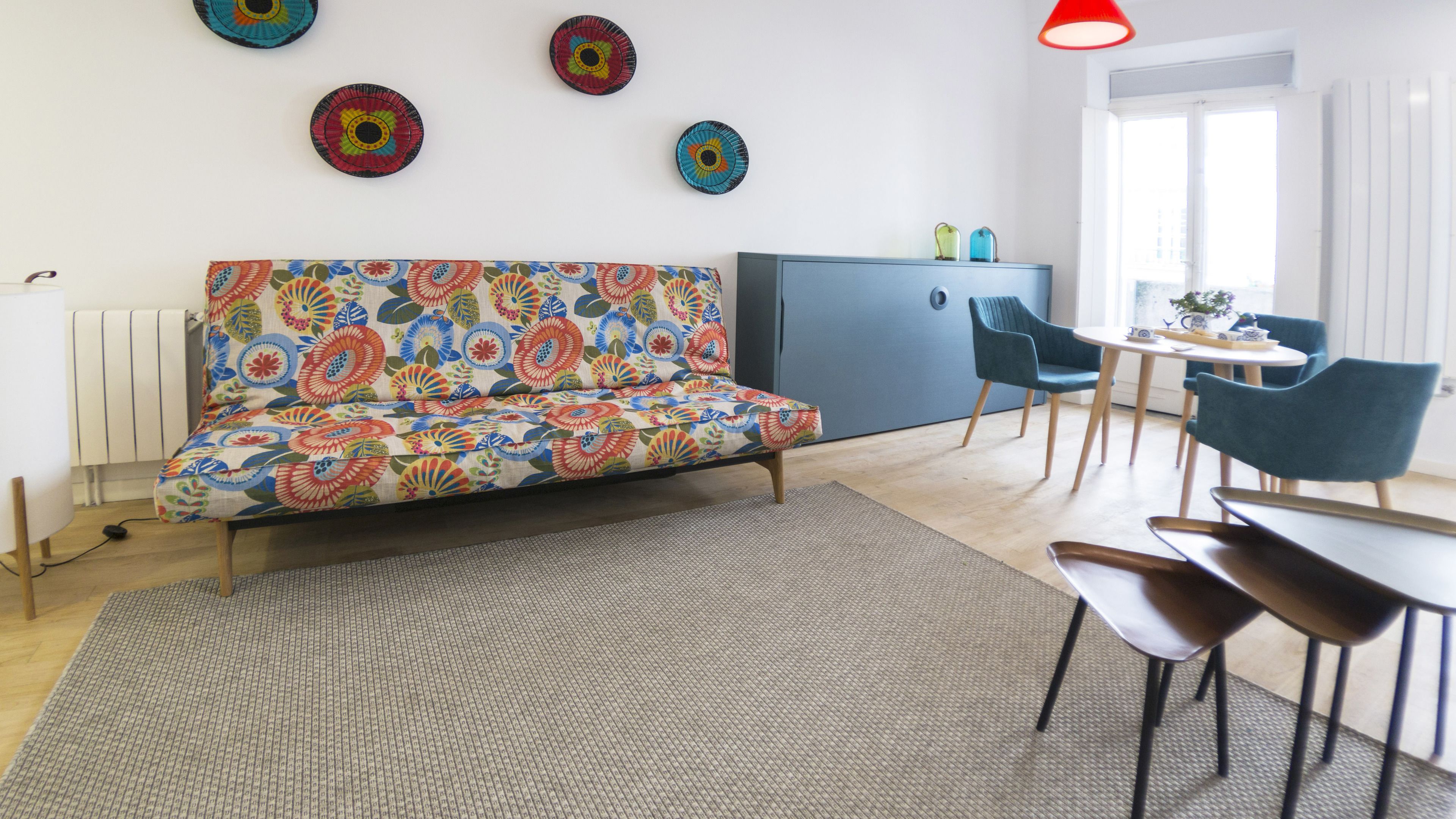 A modern living room with a colorful sofa, a dining area with chairs, a small kitchen, and decorative plates on the wall.