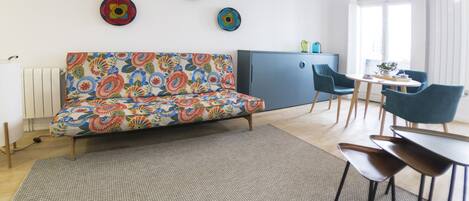 A modern living room with a colorful sofa, a dining area with chairs, a small kitchen, and decorative plates on the wall.
