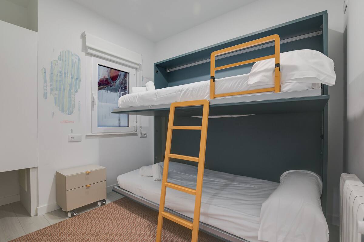 A bunk bed room with a ladder, a small cabinet, and a window with a view.