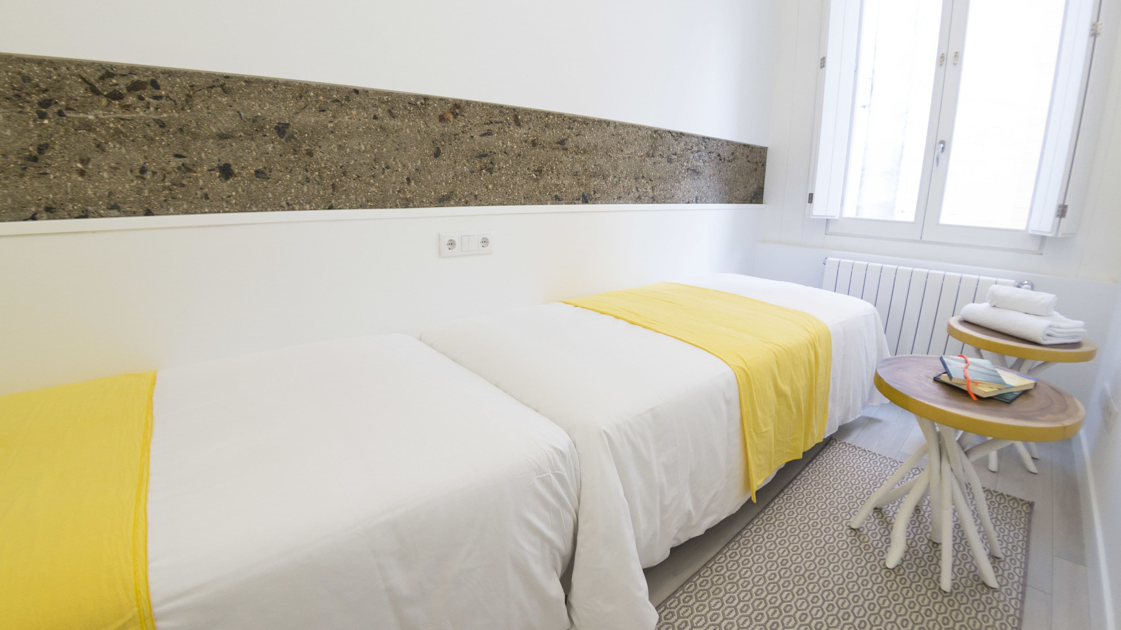 A hotel room with two beds, a small table with a book, and a radiator.