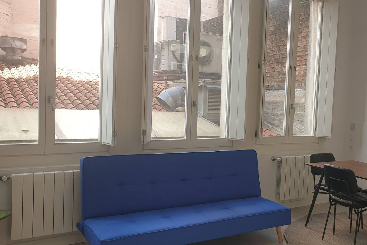 A room with a blue sofa, a wooden table with four chairs, and large windows with a view of air conditioning units outside.