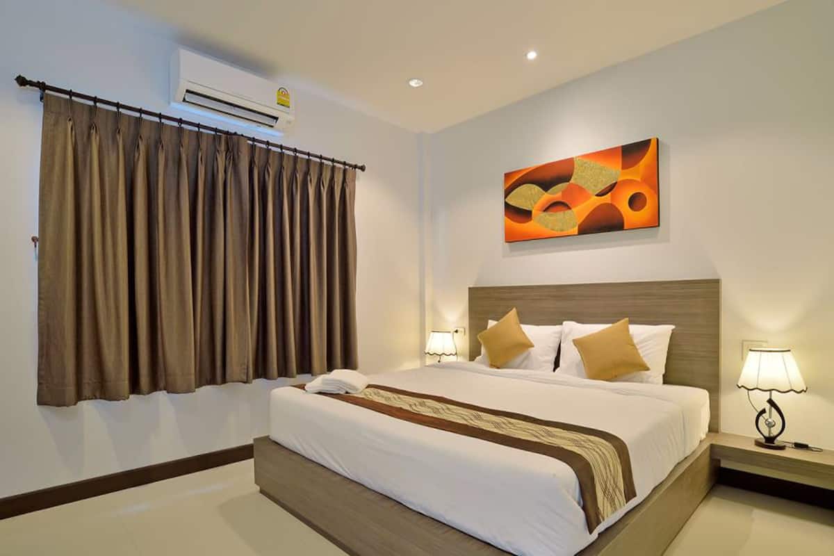 A hotel room with a large bed, a headboard, two bedside tables with lamps, a wall-mounted air conditioner, and a window with curtains.