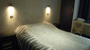 A single bed with a patterned bedspread, two wall-mounted lights, a small bedside table, and a chair by the window.