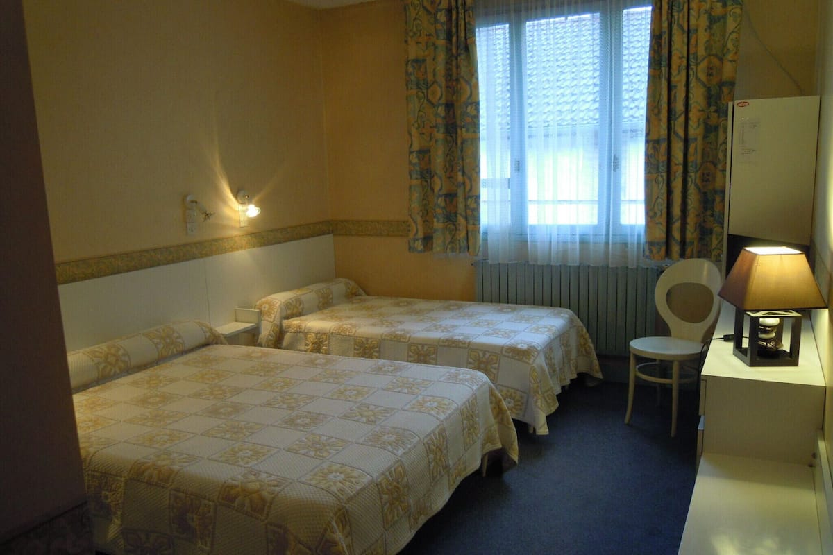 Ein Hotelzimmer mit zwei Betten, einem Schreibtisch mit Lampe, einem Stuhl und einem Fenster mit Vorhängen.