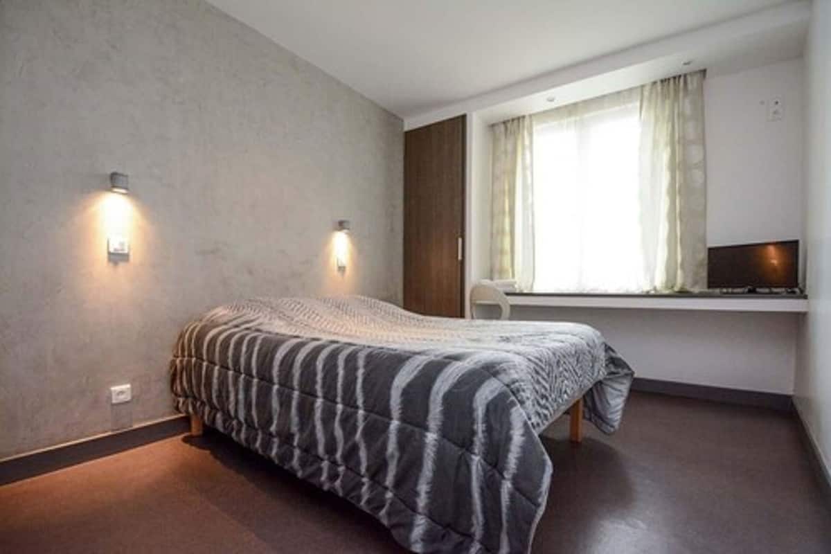 Ein Schlafzimmer mit einem Bett, zwei Wandleuchten, einem Fenster mit Vorhängen und einem Fernseher auf einem Regal.