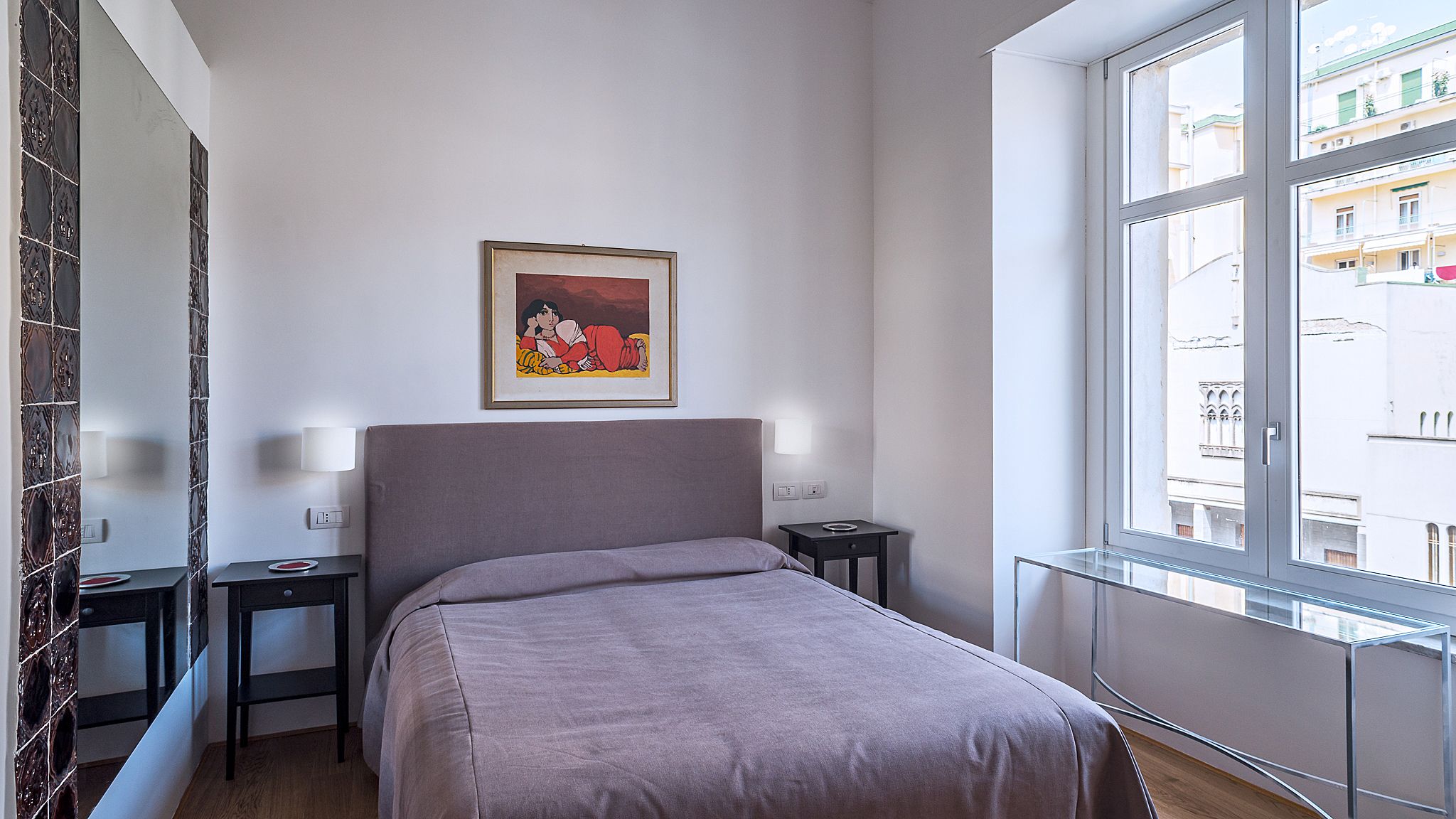 Une chambre d’hôtel avec un lit, une table de chevet, une commode, un tableau encadré au mur et une fenêtre donnant sur des immeubles.
