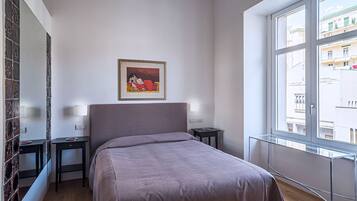 Une chambre d’hôtel avec un lit, une table de chevet, une commode, un tableau encadré au mur et une fenêtre donnant sur des immeubles.