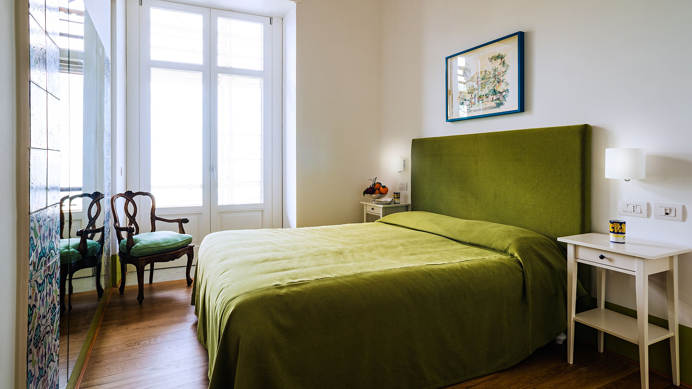 Habitación de hotel con cama, una silla, una mesita de noche con lámpara y ventana con vistas.