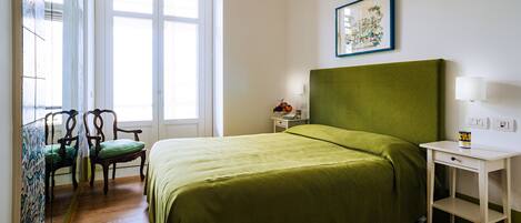 Habitación de hotel con cama, una silla, una mesita de noche con lámpara y ventana con vistas.