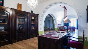A spacious library with wooden bookshelves, a central reading table, and an arched doorway leading to another room.