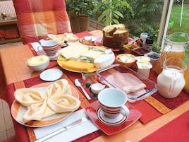 Una mesa dispuesta con diversos alimentos, incluyendo queso, jamón y una hogaza de pan, con vista a la vegetación exterior.