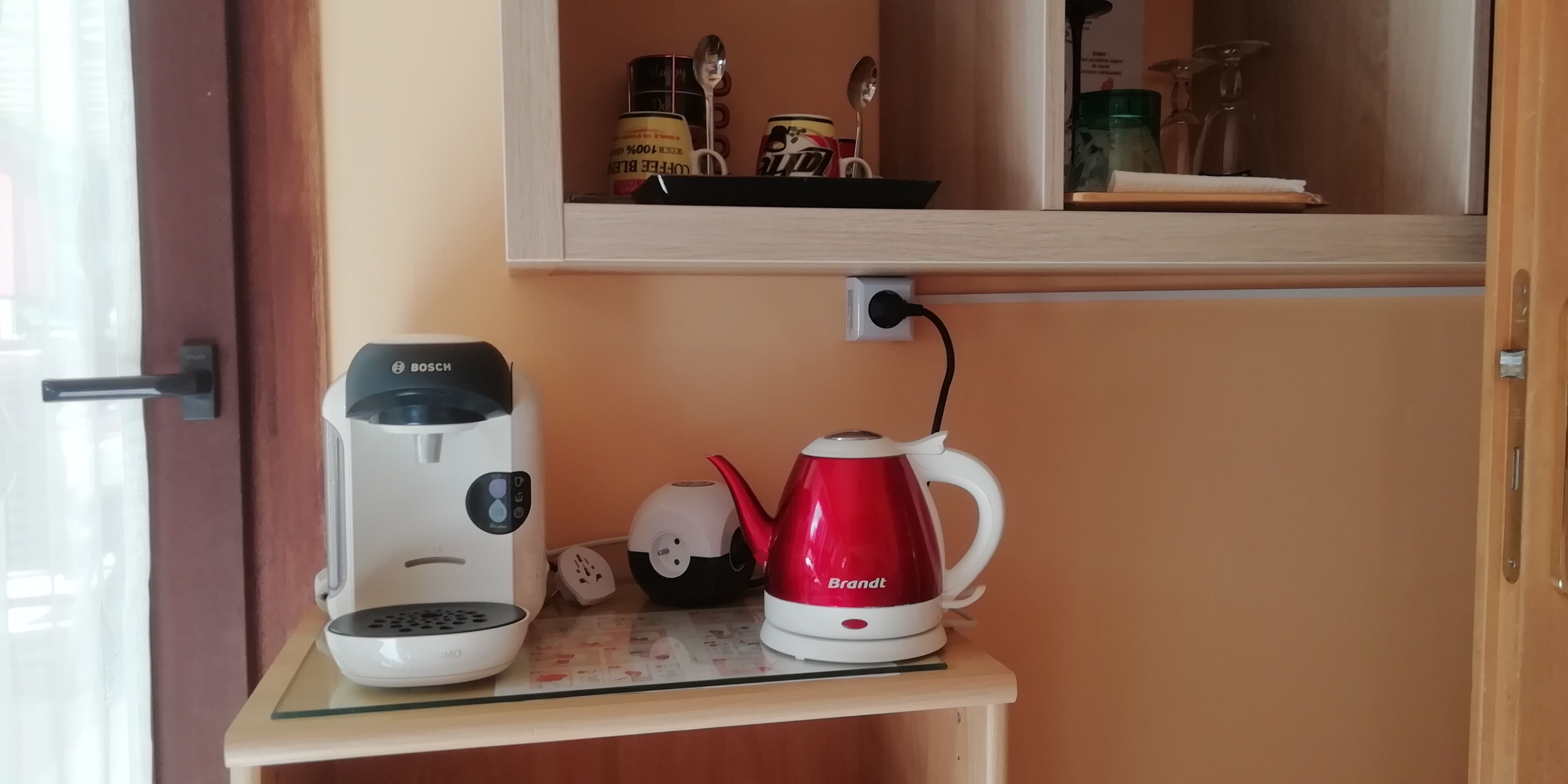 Una kitchenette con cafetera, hervidor eléctrico rojo y repisa con utensilios de cocina.