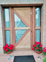 Una puerta de madera con el número 2, flanqueada por dos ventanas con cortinas y dos macetas con flores rojas en el suelo.