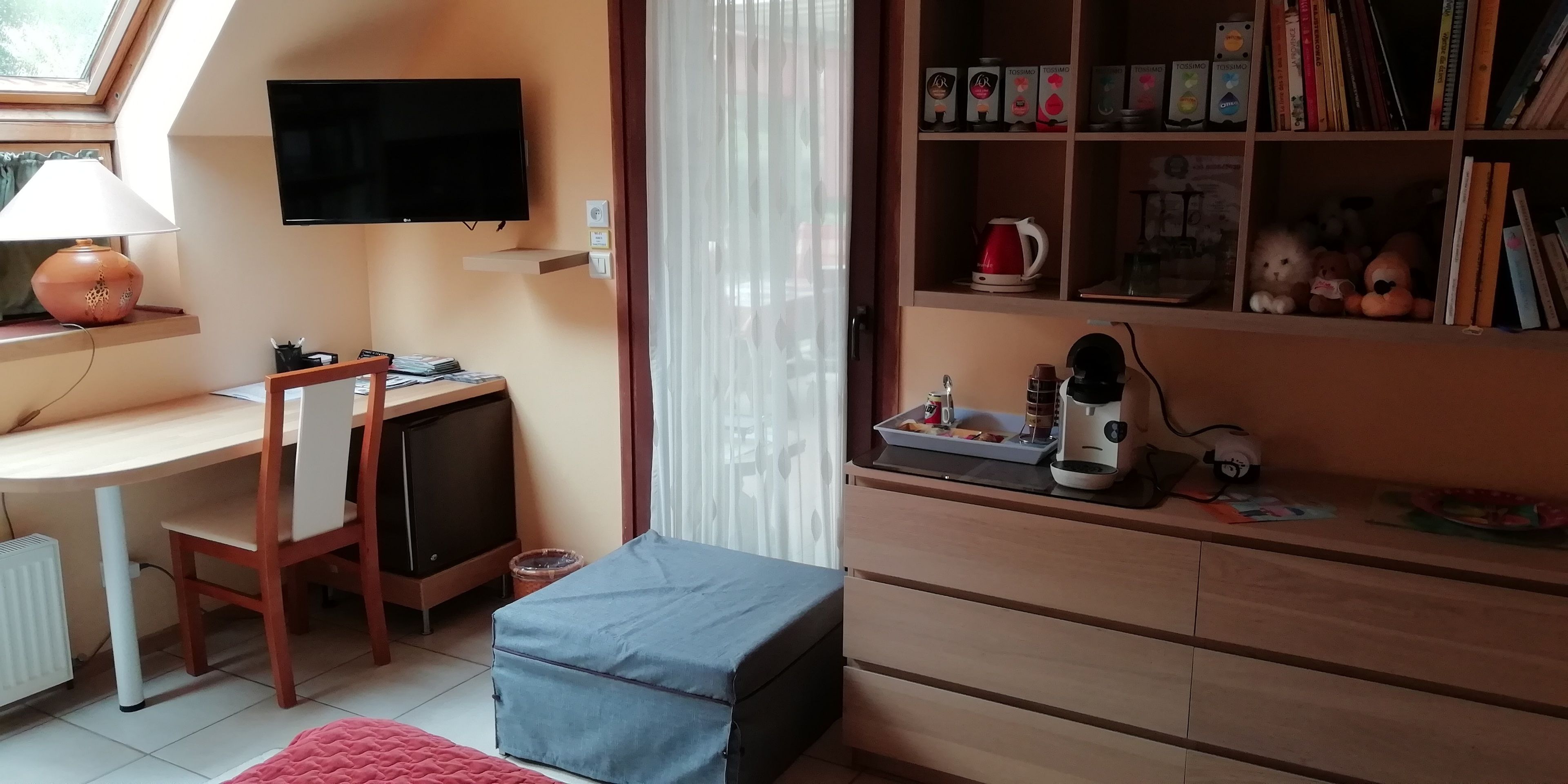 Una habitación acogedora con escritorio, silla, televisor, estantería y ventana con cortinas.