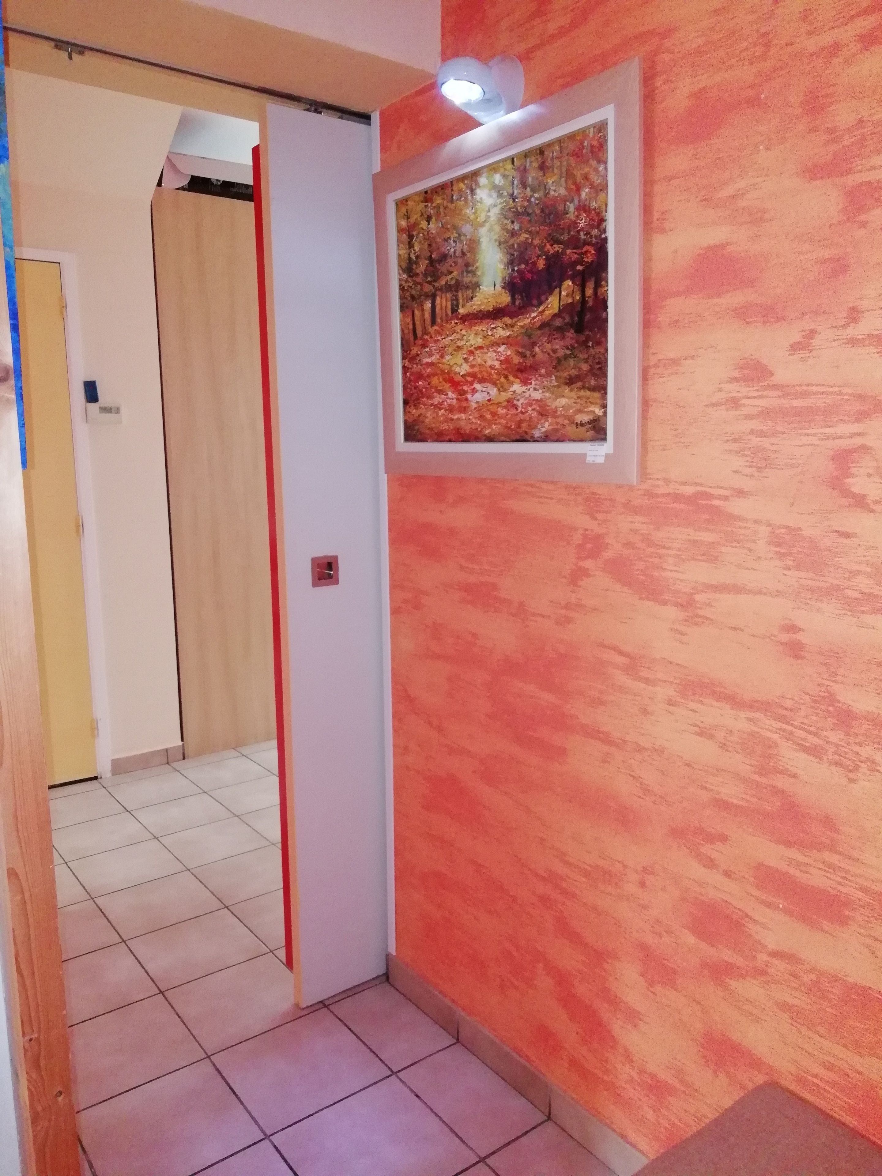 Un couloir doté d’un sol carrelé, une porte avec une bande rouge et, au mur, un tableau encadré représentant une scène de forêt.