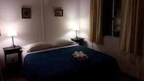 Een hotelkamer met een bed, twee bedlampen, een raam met gordijnen en twee ingelijste foto’s aan de muur.