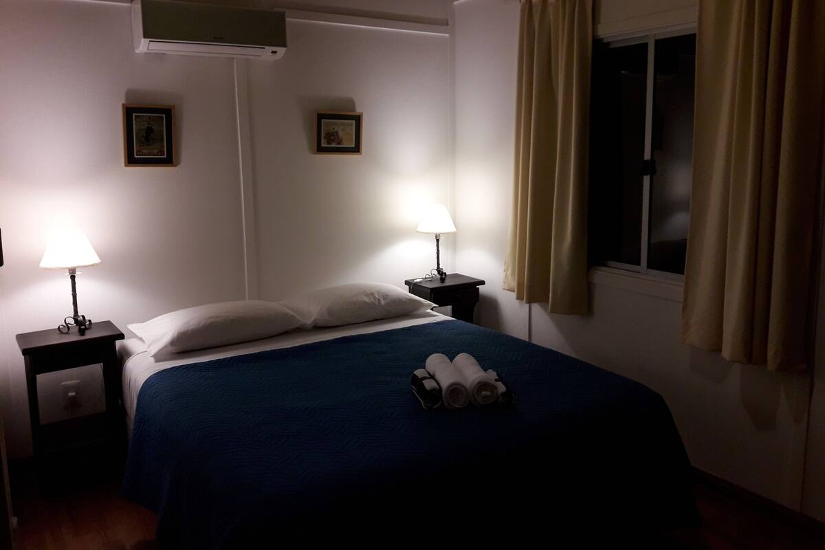 Une chambre d’hôtel avec un lit, deux lampes de chevet, une fenêtre avec des rideaux et deux tableaux encadrés accrochés au mur.