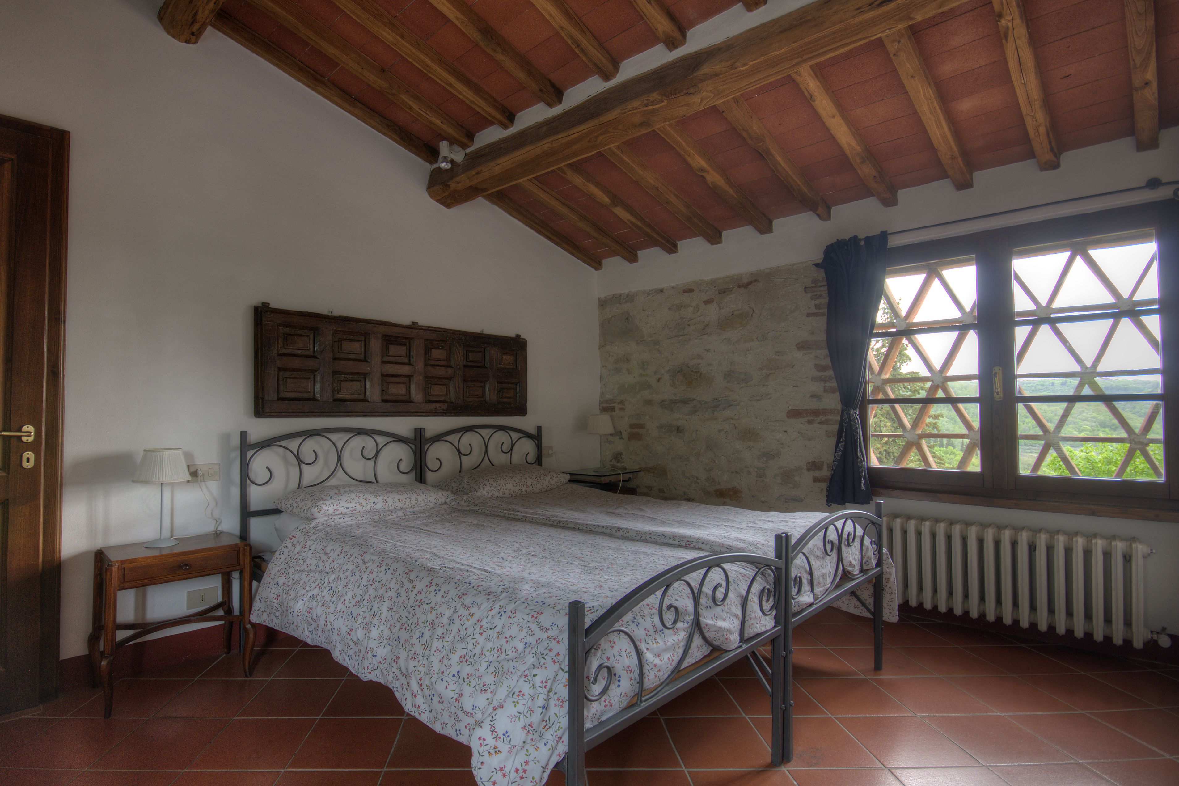 Een slaapkamer met een bed, nachtkastje, raam met uitzicht en een stenen muur.