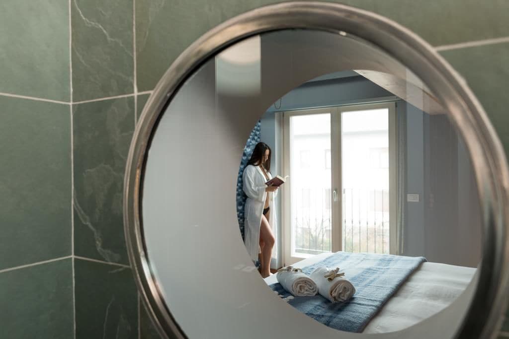 A room with a large mirror reflecting a person standing by a window.