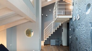 Una habitación de hotel moderna con suelo de madera, una cama con sábanas blancas, un espejo redondo, una escalera y una pared con textura.