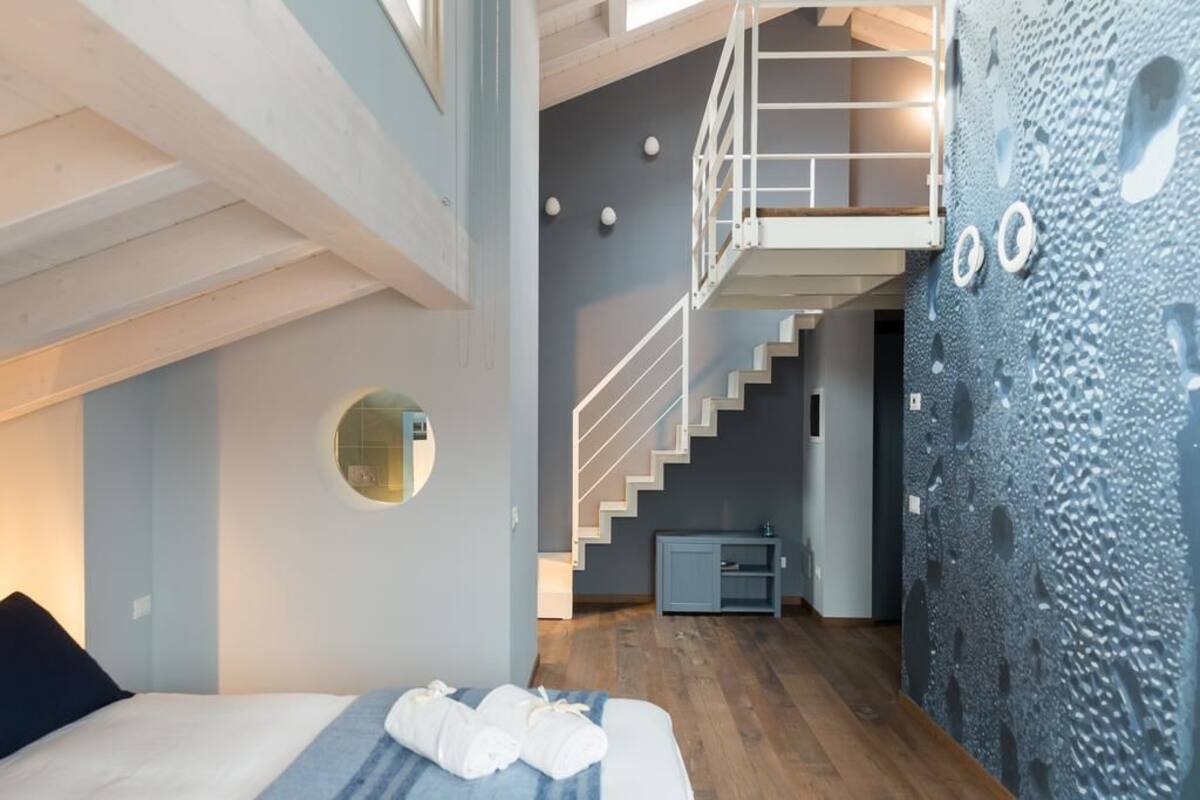A modern hotel room with a wooden floor, a bed with white linens, a round mirror, a staircase, and a textured wall.