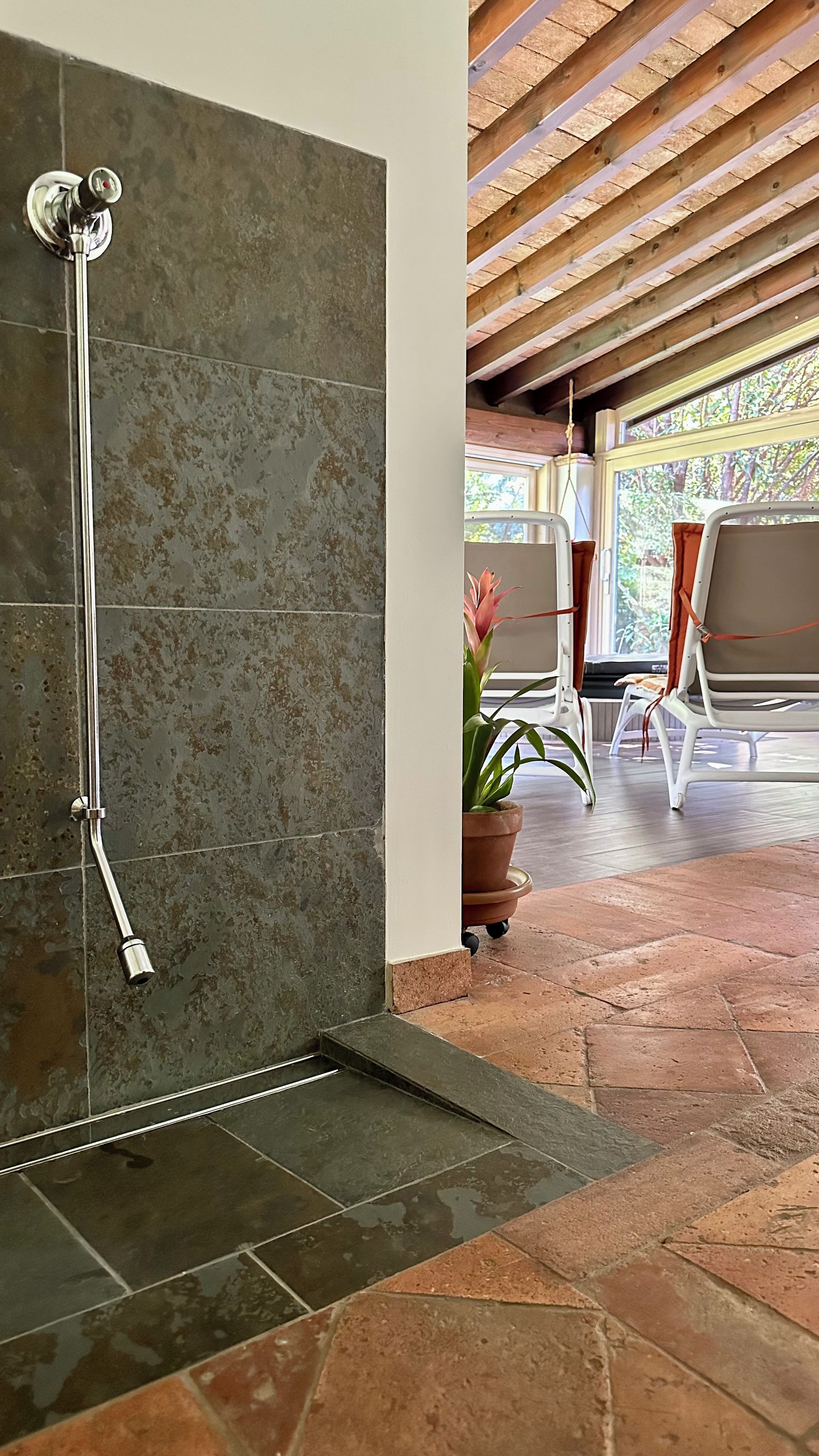 Une douche au design moderne, un plafond en bois et une vue sur un patio extérieur aménagé avec des chaises et un pot de fleurs.