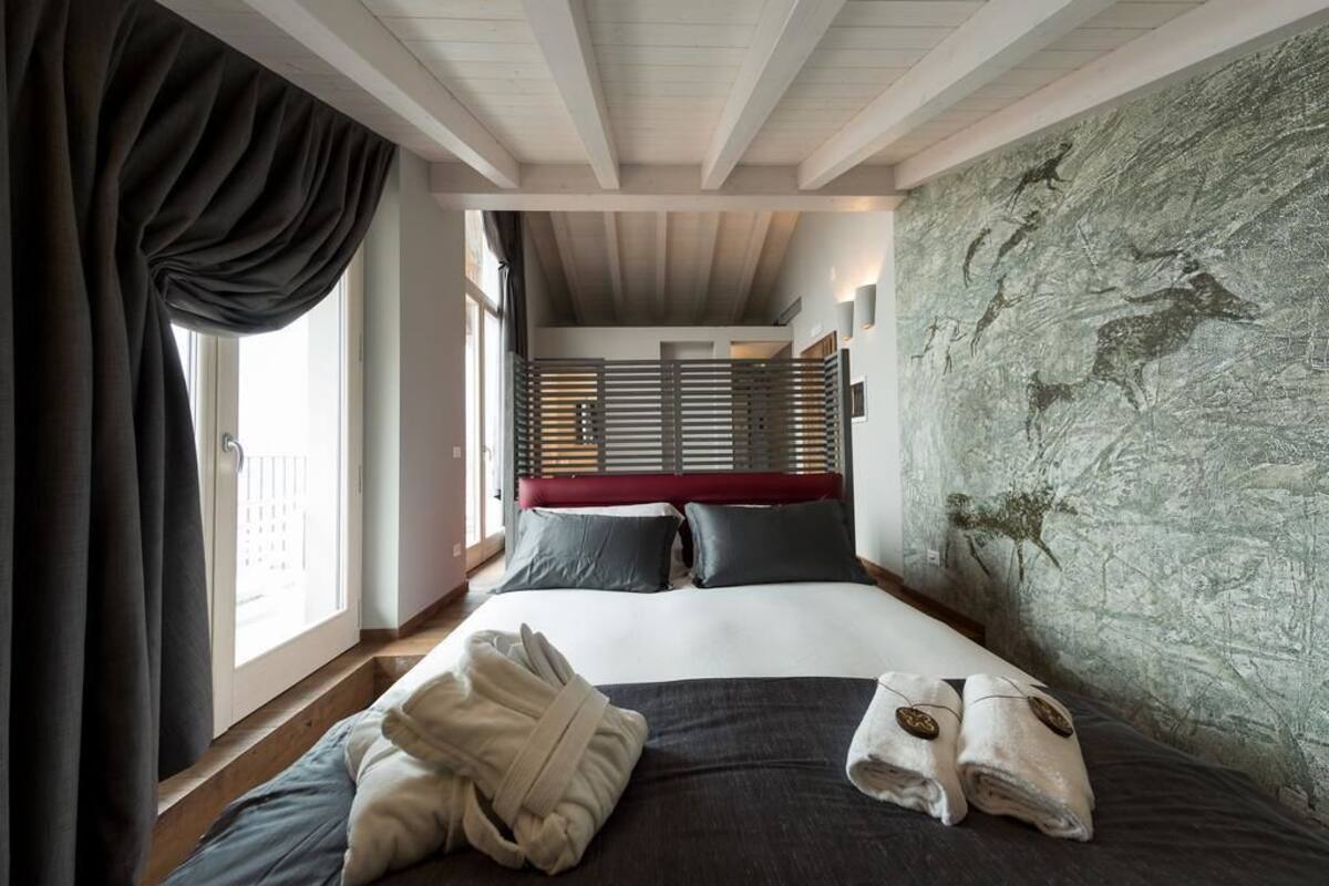 A bedroom with a bed, a window with curtains, a stone wall, and a wooden ceiling.