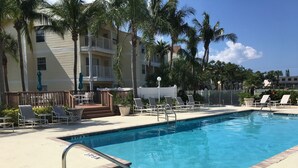Une piscine dotée de chaises longues et d’une terrasse, entourée de palmiers et bordée d’un bâtiment avec des balcons.