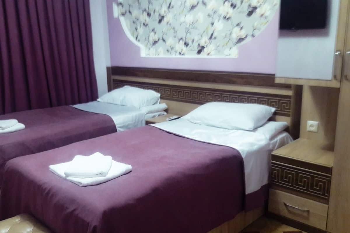 A hotel room with two single beds, each with a purple bedspread and white pillows. There is a wooden nightstand with a drawer, a wall-mounted TV, and a decorative floral wall panel.