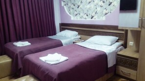 A hotel room with two single beds, each with a purple bedspread and white pillows. There is a wooden nightstand with a drawer, a wall-mounted TV, and a decorative floral wall panel. - Georgian Palace (Kutaisi)