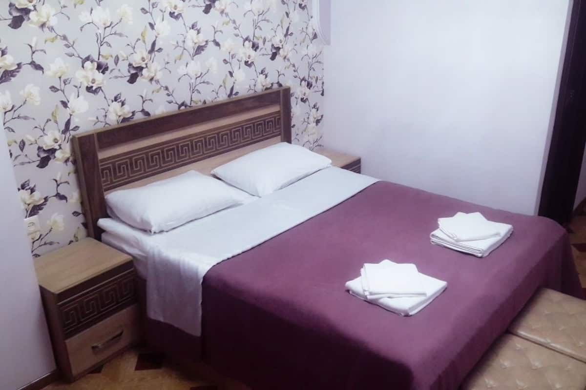 A bed with a wooden headboard, white bedding, and a purple bedspread. There is a nightstand with a drawer and a folded white towel on it. The room has a floral wallpaper and a patterned floor.