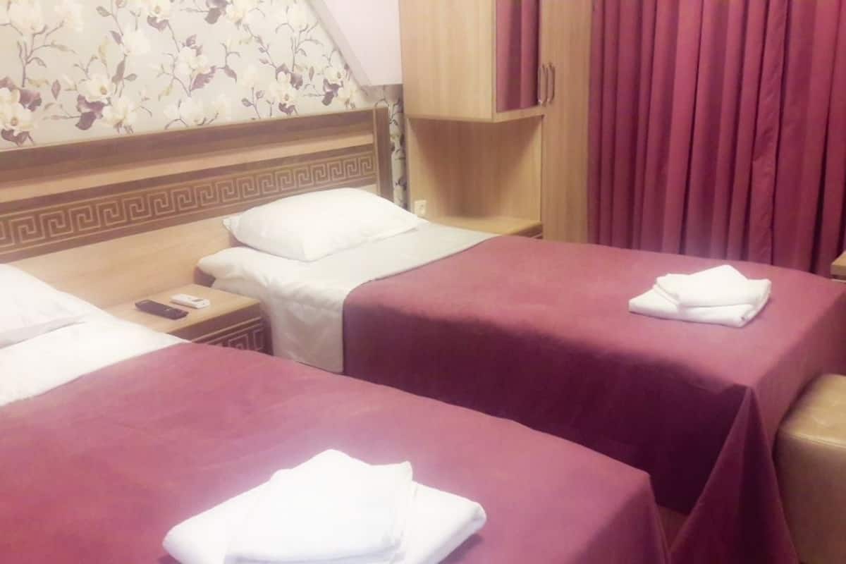 A hotel room with two beds, each with a red bedspread and white pillows. There is a wooden headboard with a decorative pattern, a wooden nightstand with a phone and a remote control, and a wooden cabinet above the beds.