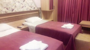 A hotel room with two beds, each with a red bedspread and white pillows. There is a wooden headboard with a decorative pattern, a wooden nightstand with a phone and a remote control, and a wooden cabinet above the beds. - Georgian Palace (Kutaisi)
