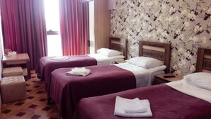 A hotel room with three beds, each with a wooden headboard and white bedding. There is a small table with a lamp and a chair, and a window with curtains. - Georgian Palace (Kutaisi)