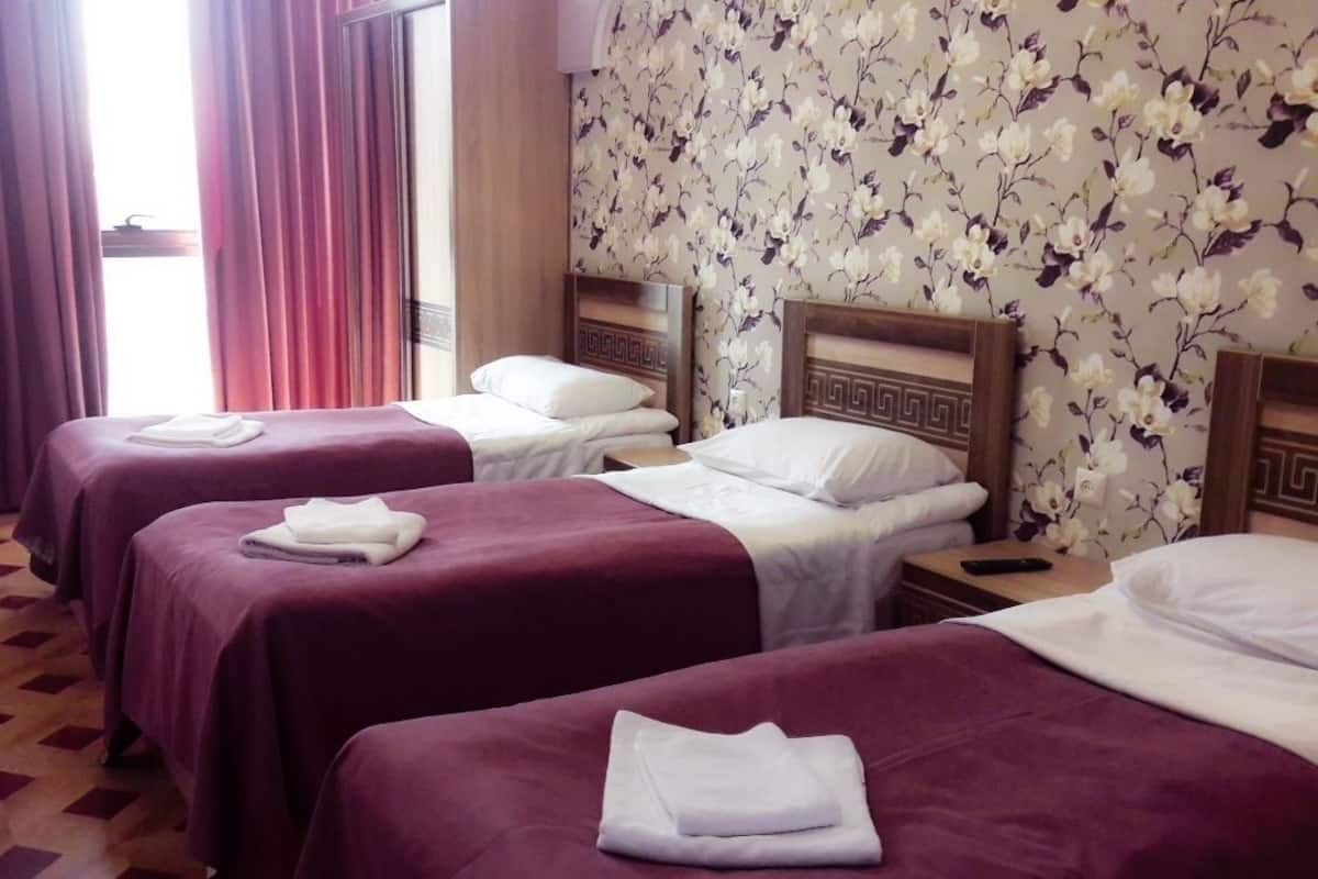 A hotel room with three beds, each with a wooden headboard and white bedding. There is a small table with a lamp and a chair, and a window with curtains.