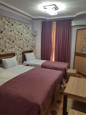 Habitación de hotel con dos camas, un armario de madera, una mesita pequeña y un papel tapiz con motivos florales.