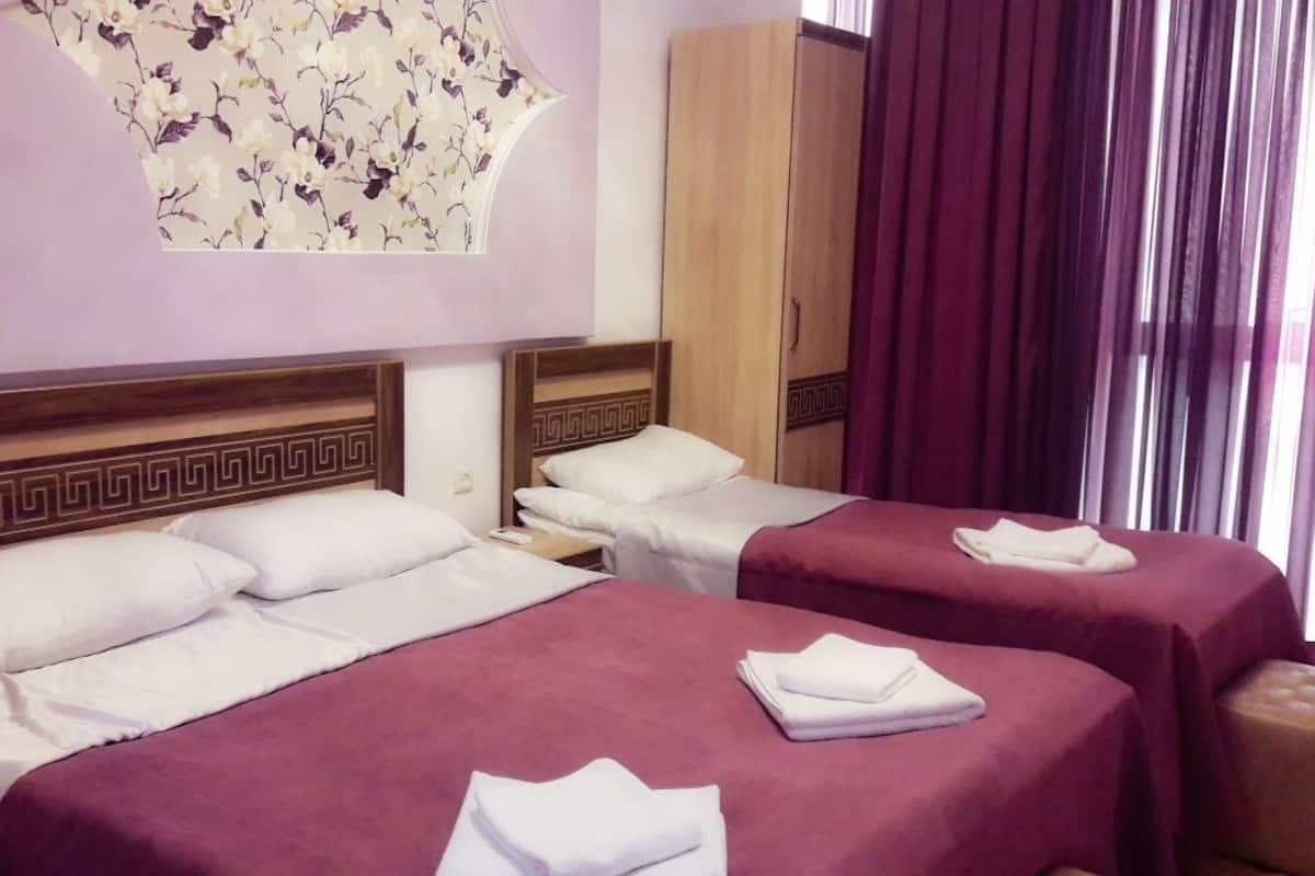 A hotel room with two beds, each with a purple bedspread and white pillows. There is a wooden headboard with a decorative pattern, a wardrobe, and a large floral artwork on the wall.