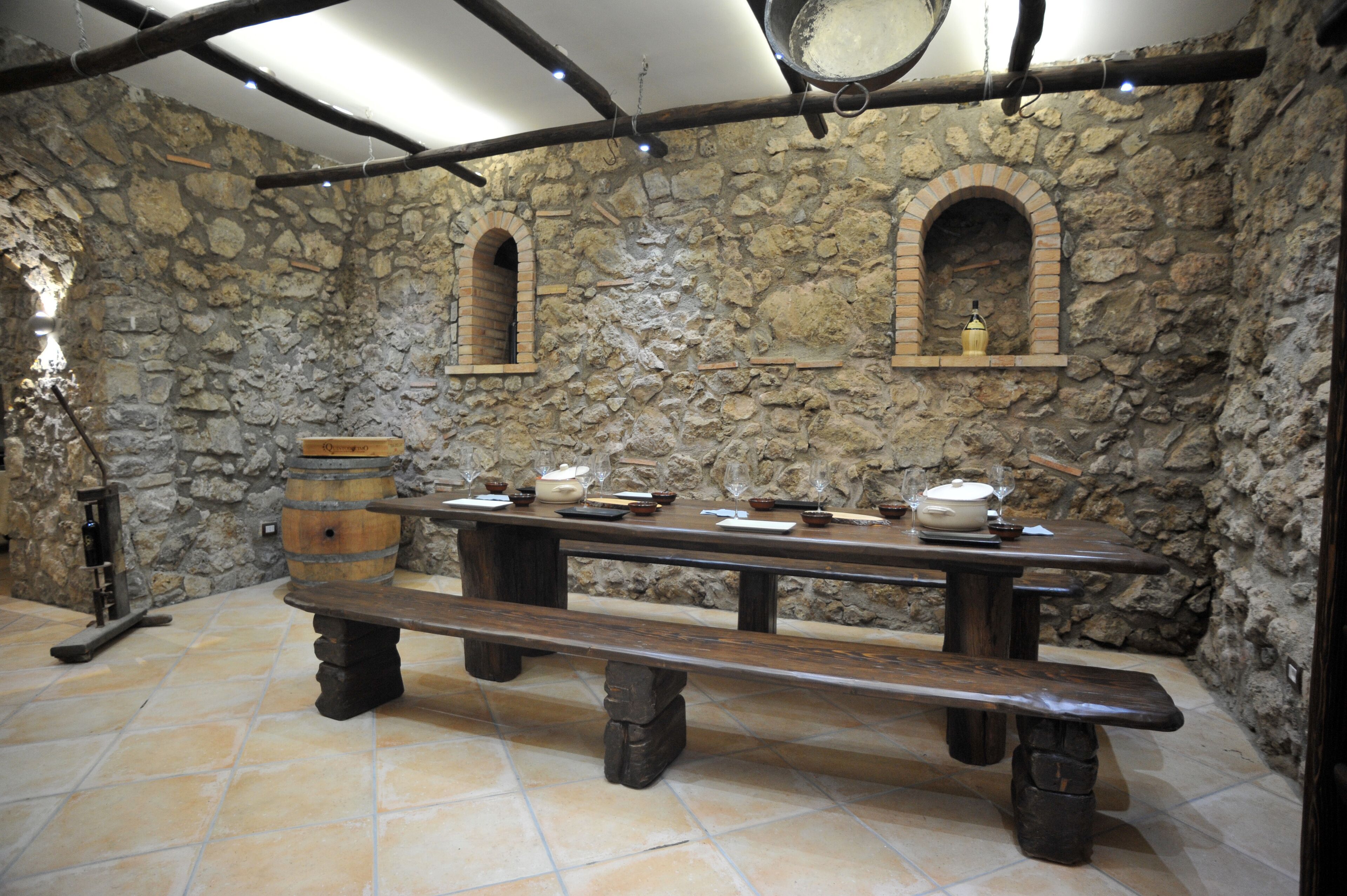 A stone-walled room with a long wooden table set for dining, two arched niches with pottery, and a hanging pot.