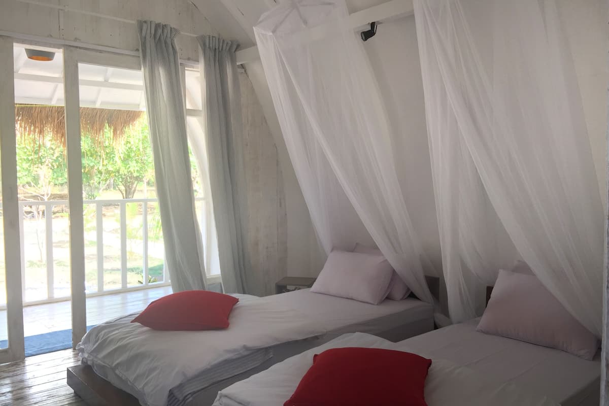 Habitación con dos camas, cada una con ropa blanca y almohadas rojas. Se ve un ventanal con cortinas y un balcón al exterior.