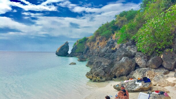 A beach with clear turquoise water, rocky cliffs, and a group of people relaxing on a towel. - Maleo Moyo Seaside Resort (Moyo)