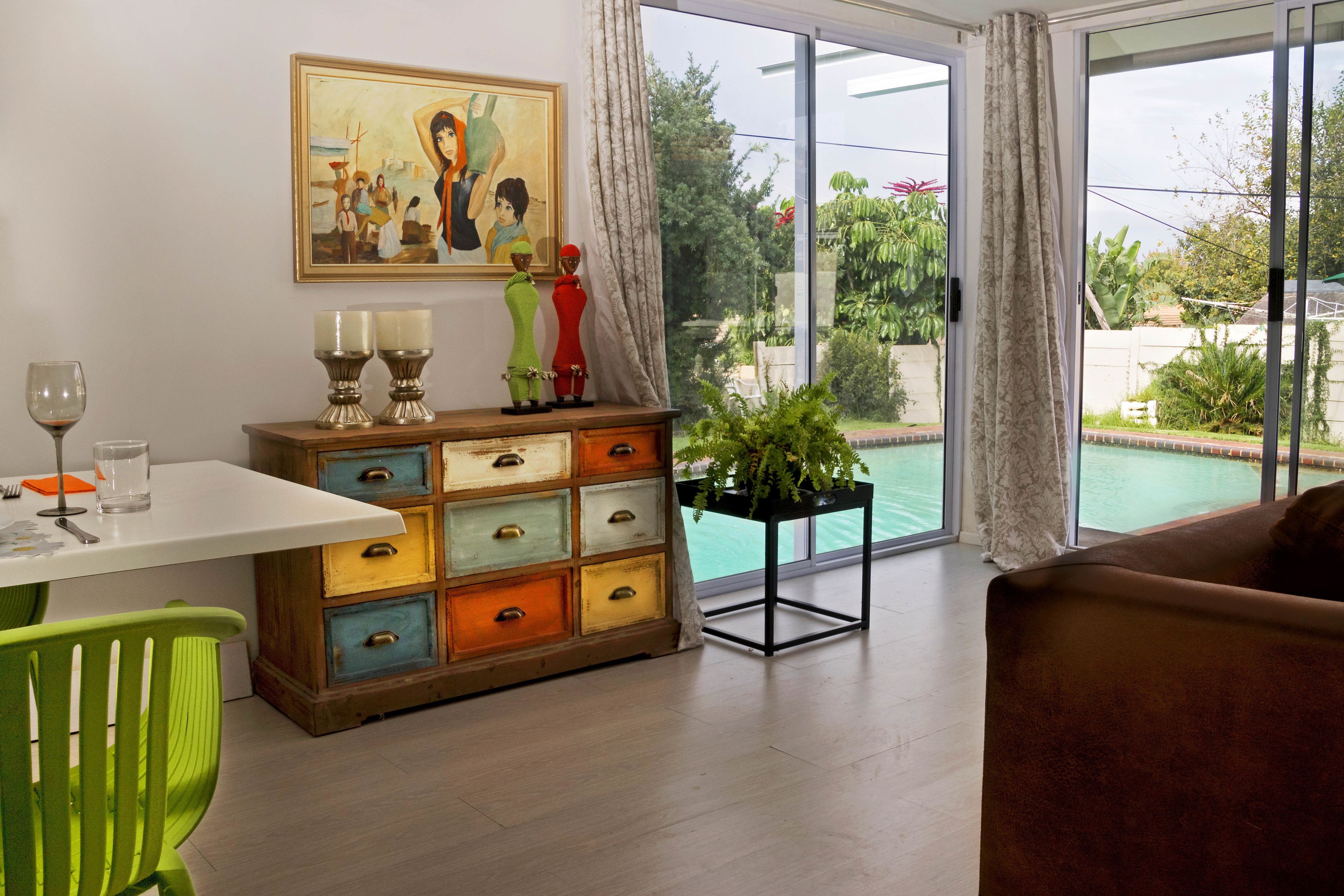 A dining area with a table, chairs, a colorful chest of drawers, a painting, and a view of a swimming pool through large windows.