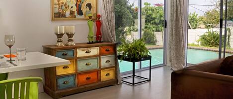 A dining area with a table, chairs, a colorful chest of drawers, a painting, and a view of a swimming pool through large windows.