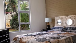 Une chambre avec une grande fenêtre, un lit recouvert d’une couvre-lit à motifs, une table de chevet avec une lampe et une tête de lit décorative.