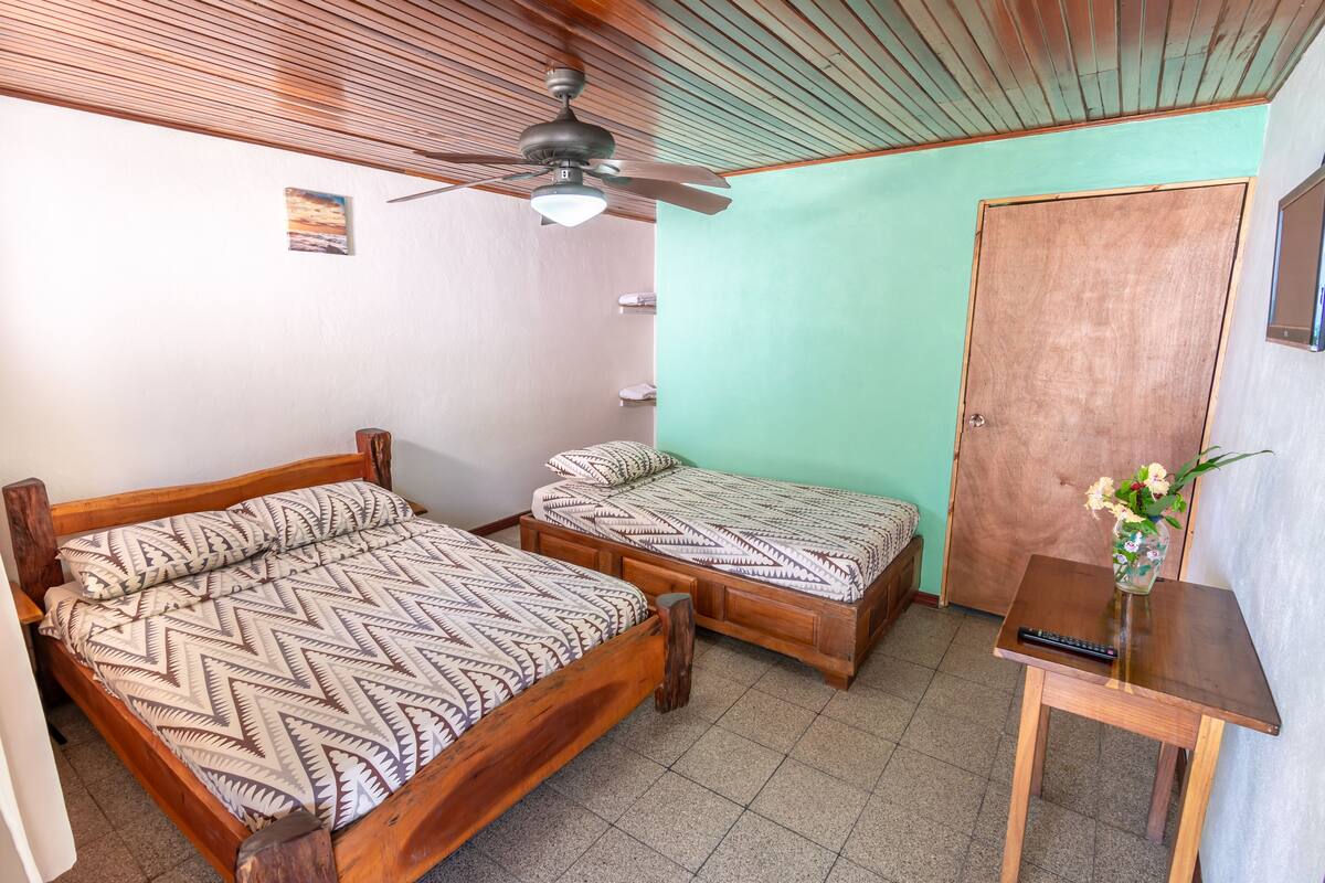 A room with two wooden beds, a ceiling fan, a small table with a vase, and a wooden door.